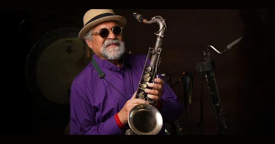 Foto oficial de Bozar para Coltrane 100: el saxofonista Joe Lovano, con sombrero y camisa morada, sostiene su saxofón tenor sobre fondo oscuro.